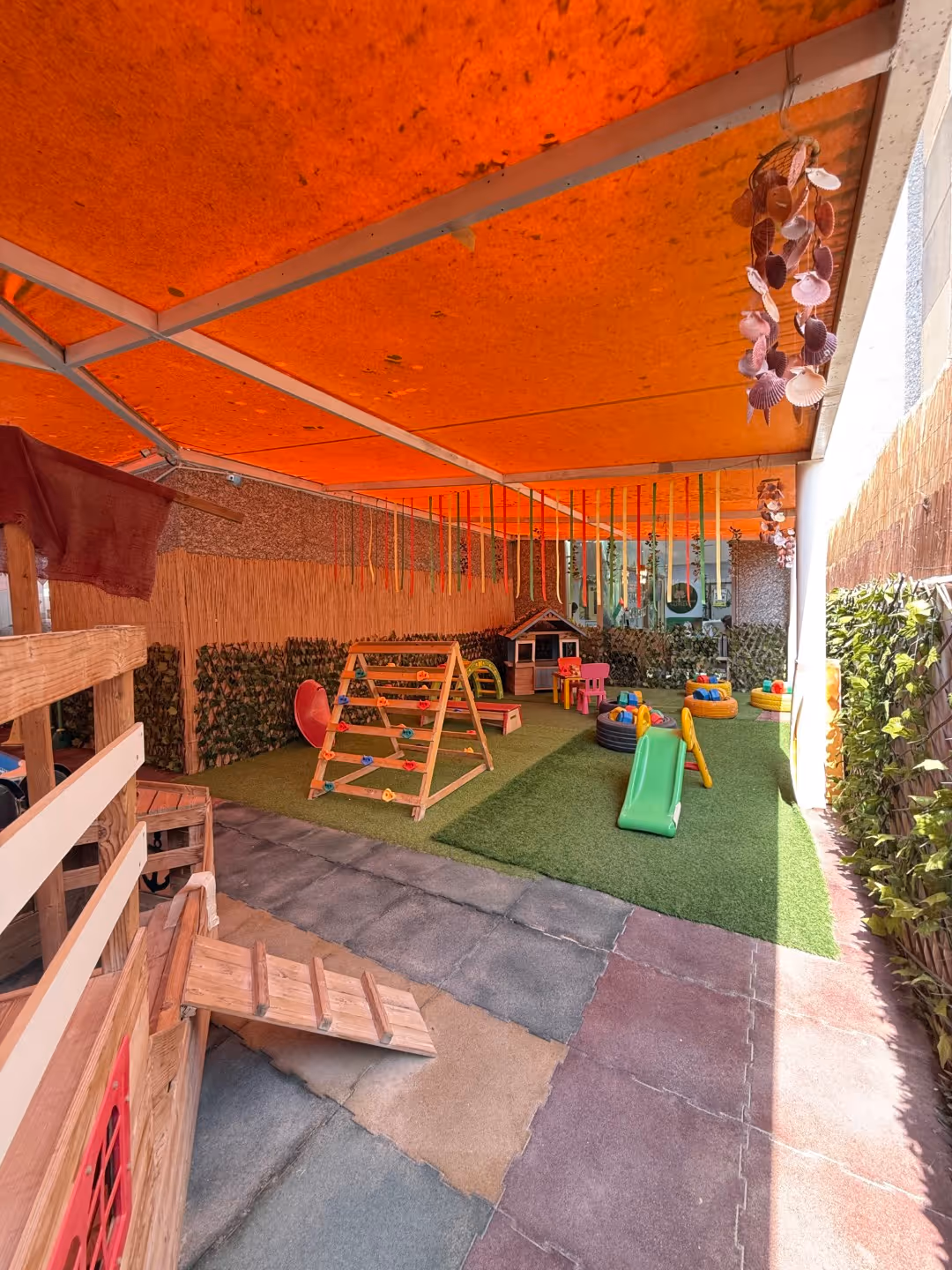 Covered outdoor children's play area with climbing structures, slide, small tables, and artificial grass under orange canopy.