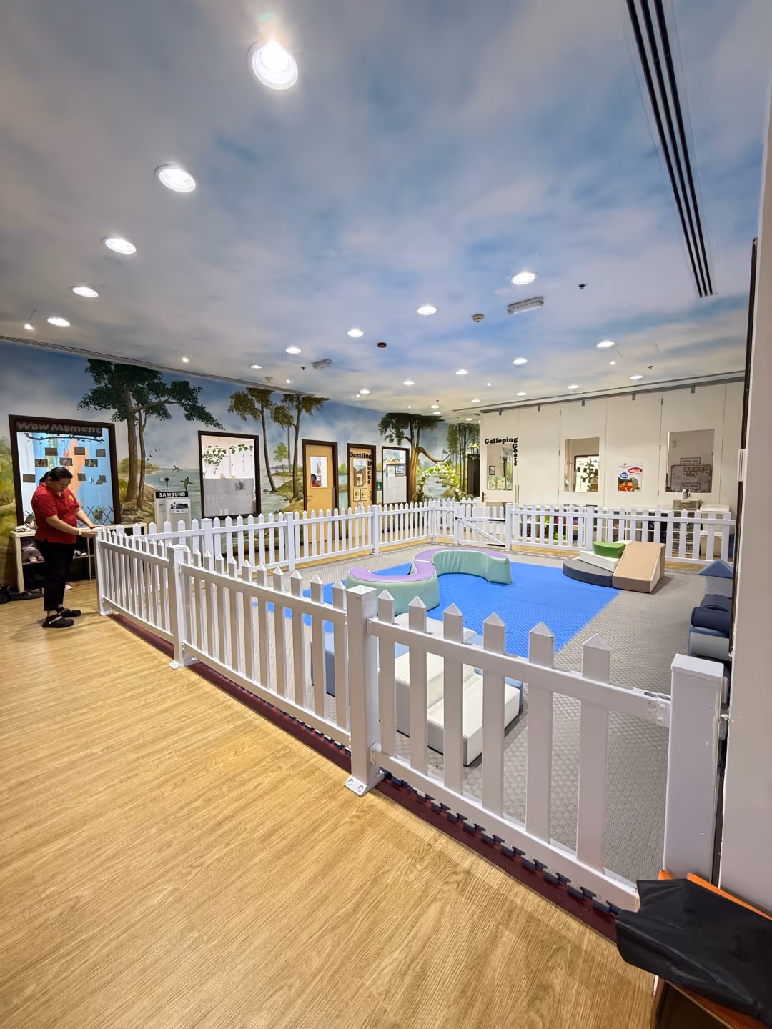 Indoor play area enclosed by white picket fences with colorful cushioned seating and murals of trees on the walls.