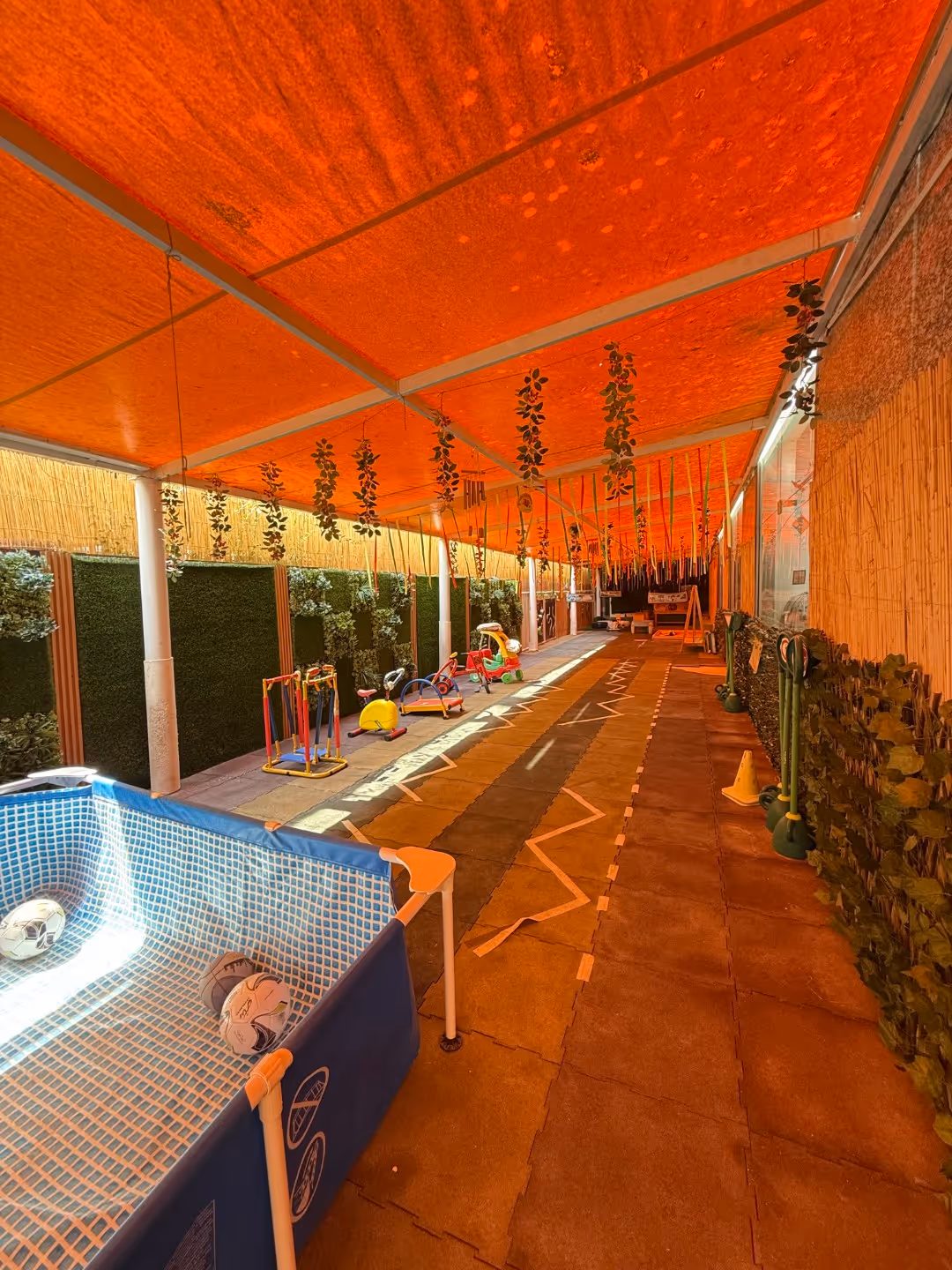 Covered indoor play area with hanging plants, small play equipment, inflatable pool with soccer balls, and foam floor mats.