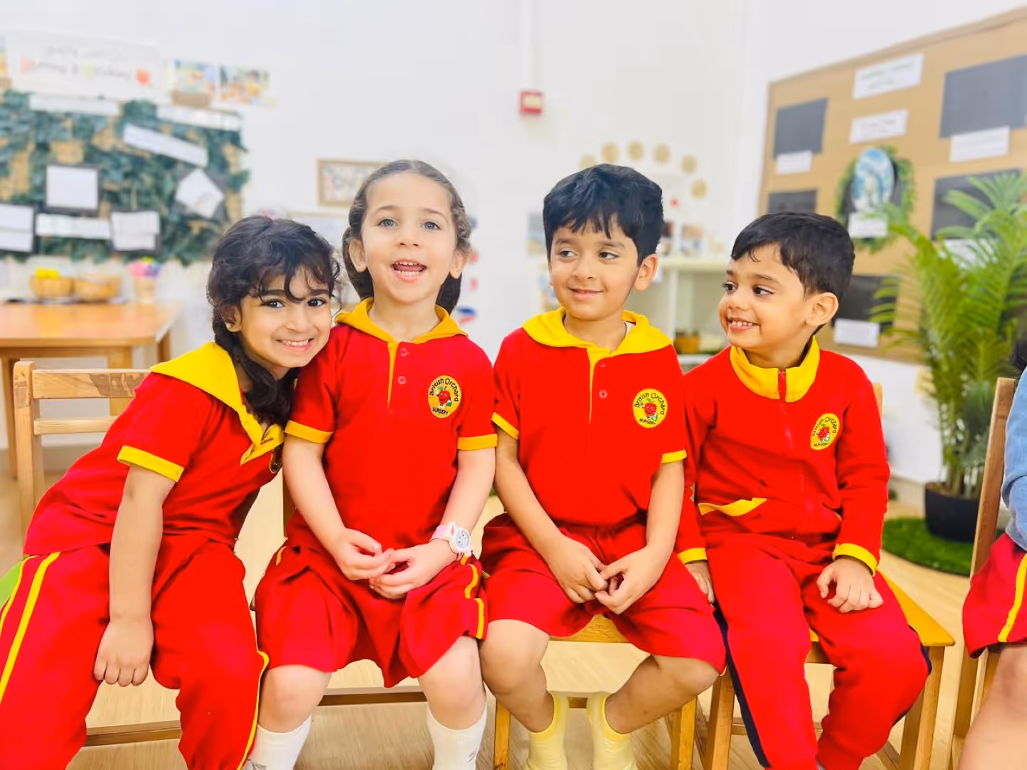 Four children in red and yellow school uniforms sitting closely on chairs in a classroom, smiling and interacting.