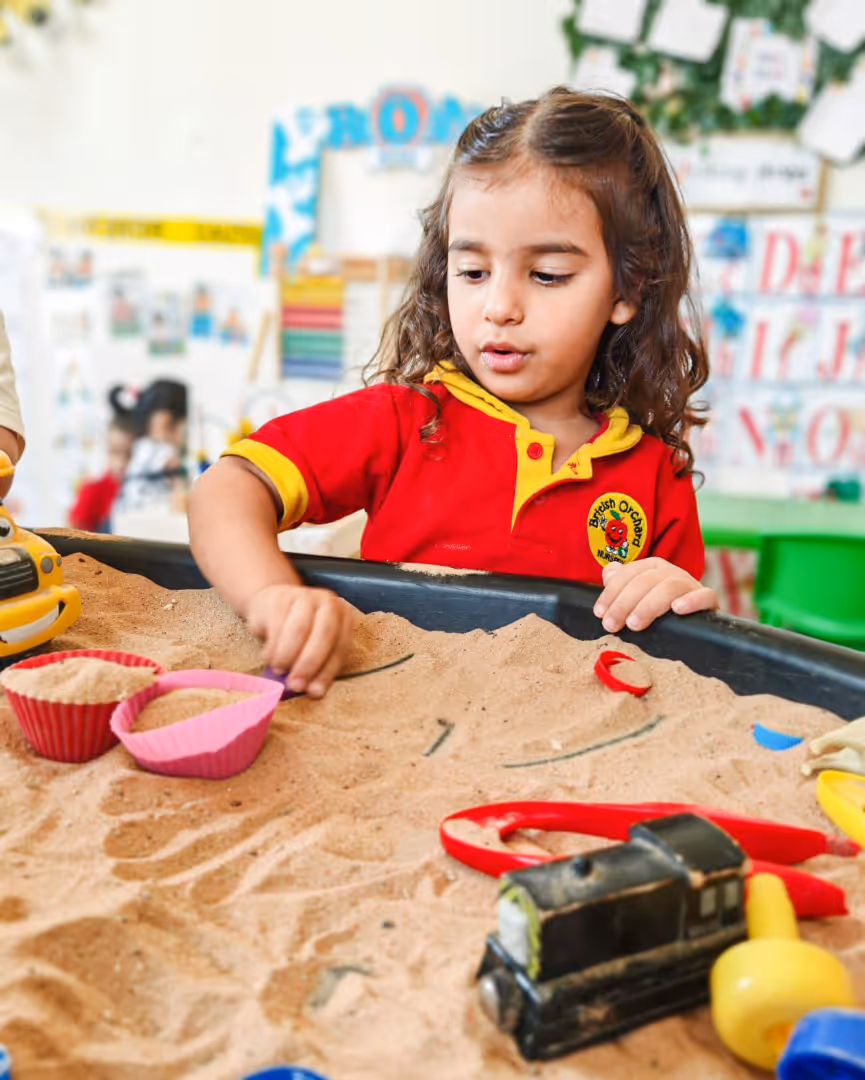 Young girl in a red and yellow shirt playing with sand and toys in a classroom setting.