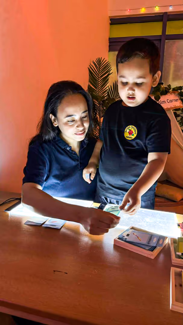 A woman and a young boy looking at illuminated cards on a table in a warmly lit room with plants and string lights in the background.