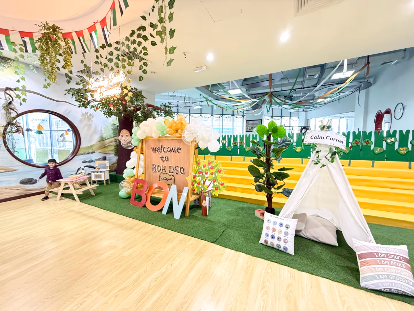 Bright indoor play area with a welcome sign, balloons, a small tent labeled Calm Corner, colorful cushions, and a child near a picnic table.