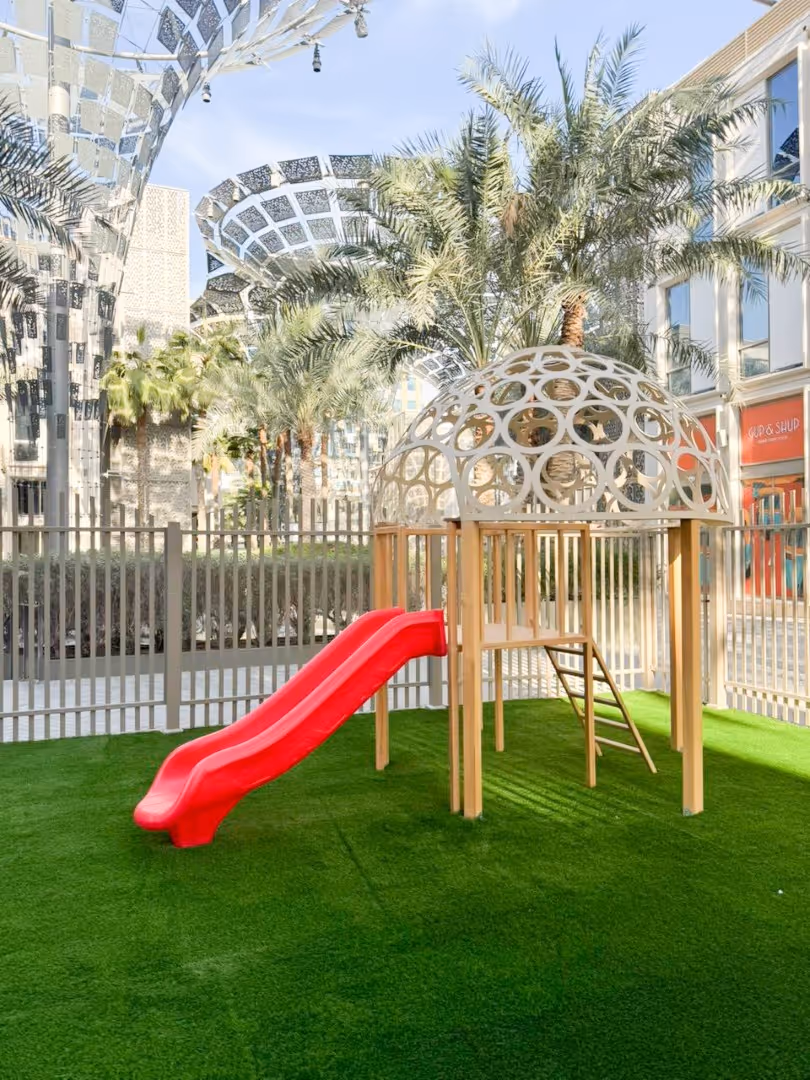 Outdoor playground with a red slide and a beige circular climbing structure on green artificial grass surrounded by palm trees and modern buildings.