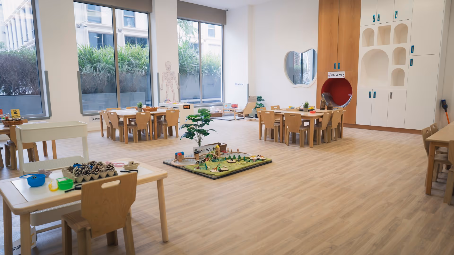 Bright and spacious preschool classroom with wooden tables and chairs, educational toys, and large windows letting in natural light.