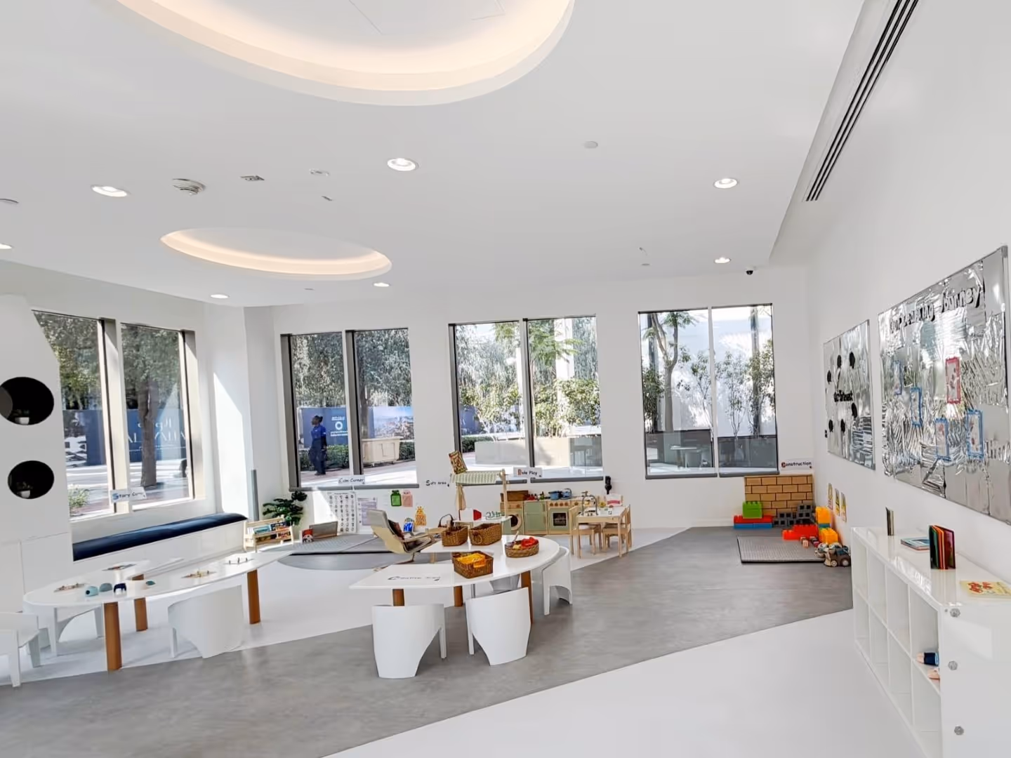 Bright and modern preschool classroom with low tables, chairs, educational toys, and large windows showing outdoor greenery.