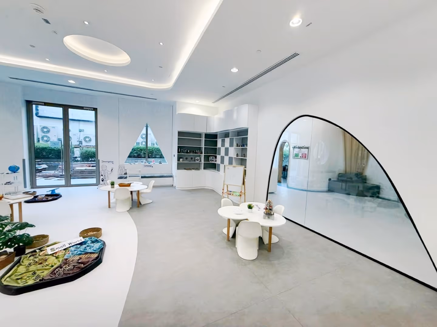 Bright, modern classroom with round tables, small white chairs, shelves, and a large curved window.