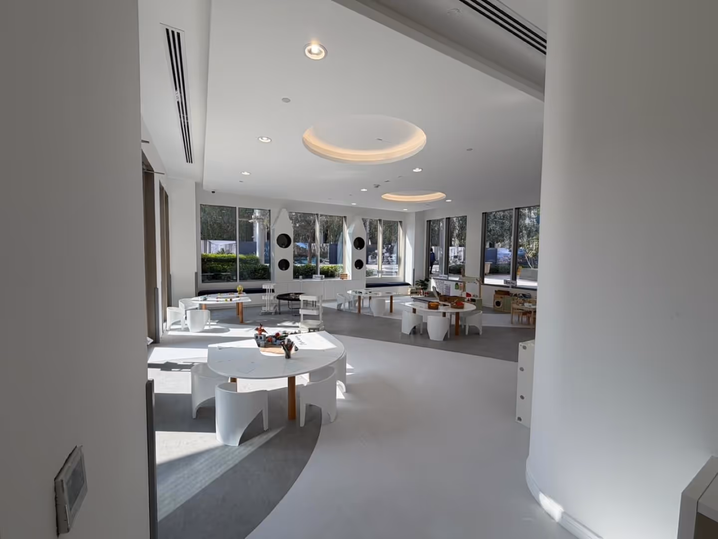 Bright, modern classroom with round white tables and small chairs arranged for children, illuminated by large windows.