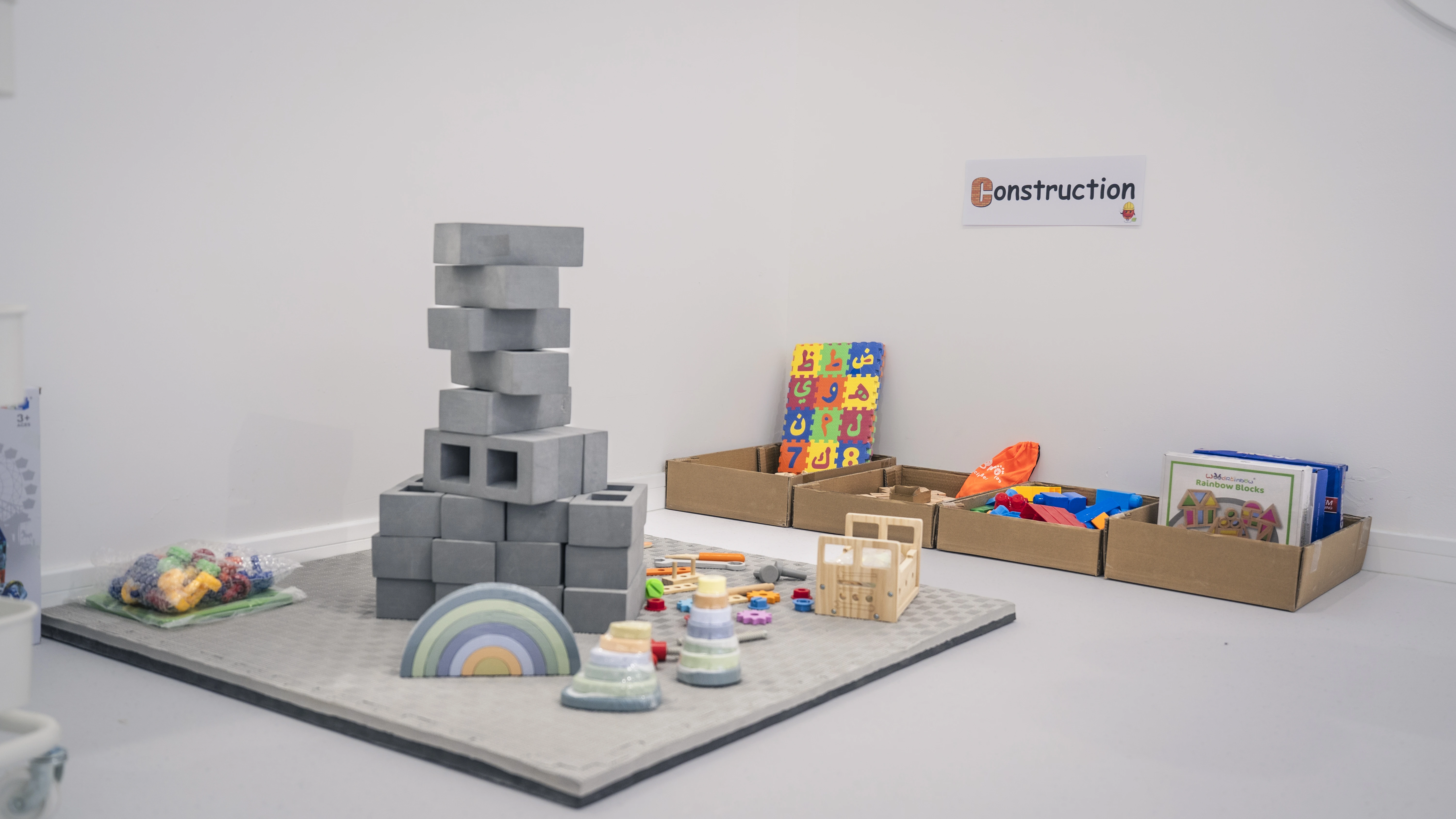 Children's play area with foam building blocks stacked on a grey mat, wooden toys, and colorful educational materials in cardboard trays under a sign that says Construction.