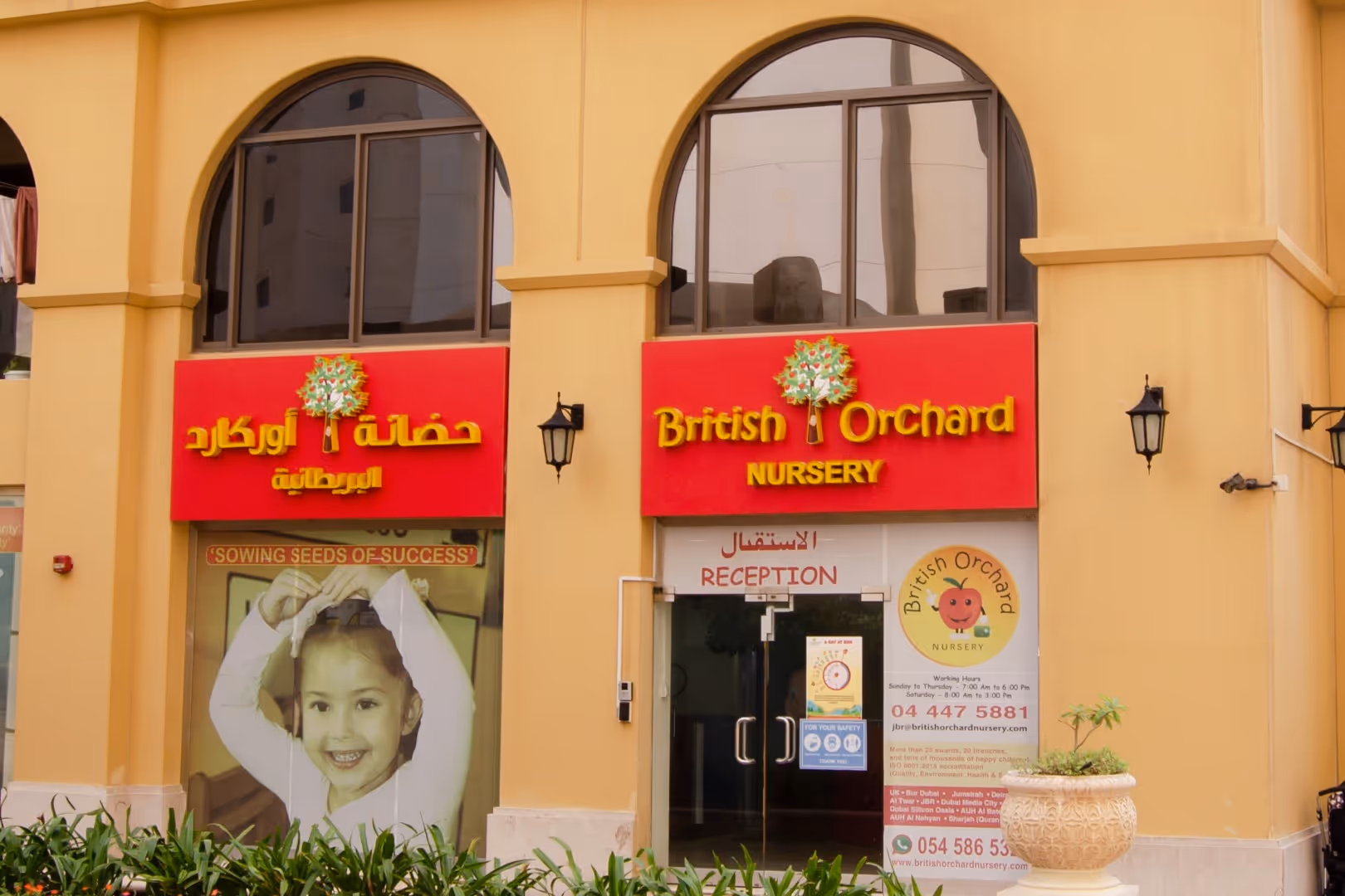Entrance of British Orchard Nursery with red signs in English and Arabic, a large photo of a smiling child, and plants in front.