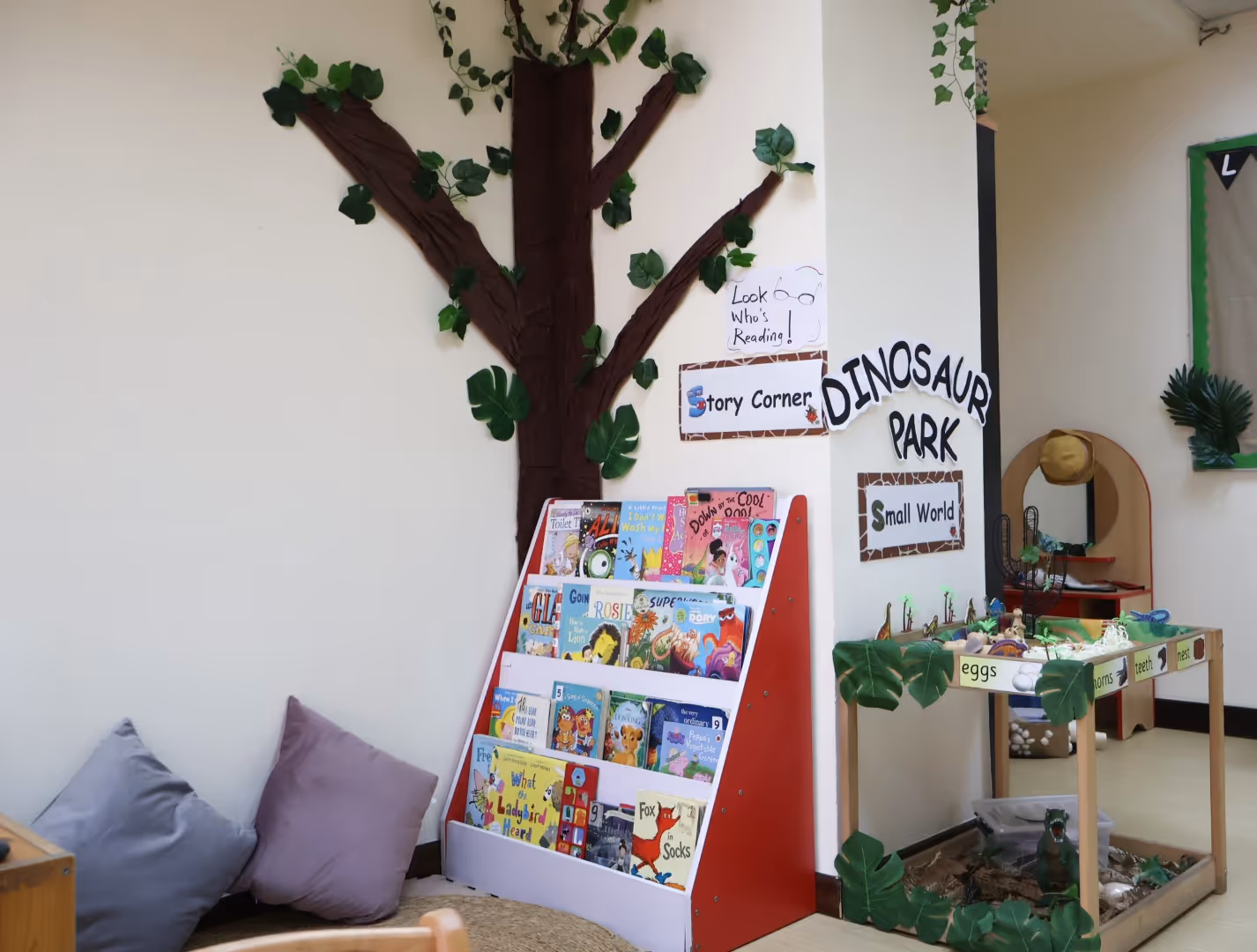 Children's reading corner with a book rack, a decorative tree on the wall, floor cushions, and a small dinosaur park play area with toy dinosaurs.