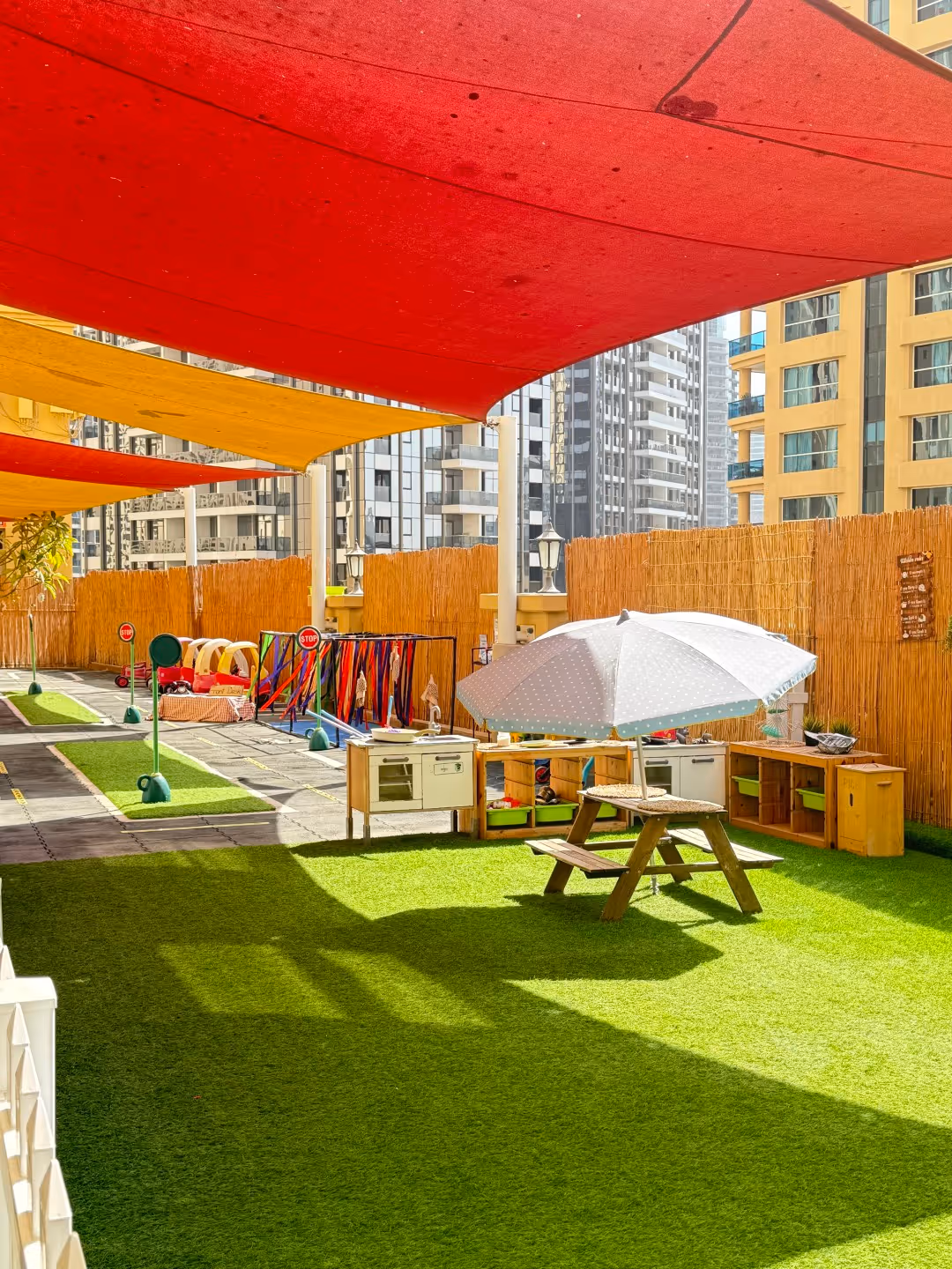 Outdoor children's play area with artificial grass, wooden picnic table under a white umbrella, colorful shade sails overhead, and nearby toys and storage shelves.