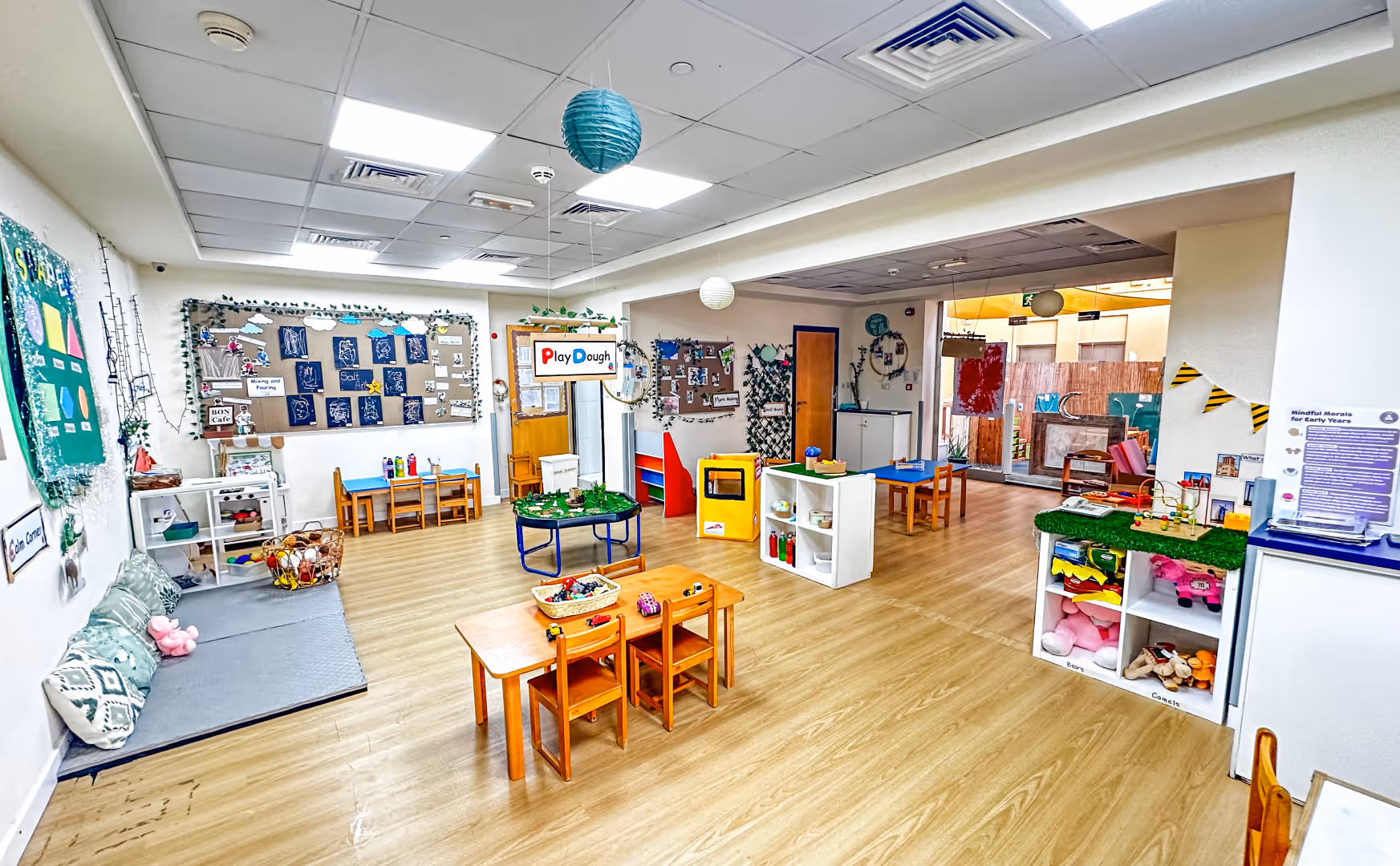 Bright and organized preschool classroom with small wooden tables and chairs, educational posters, play areas, and colorful toys.