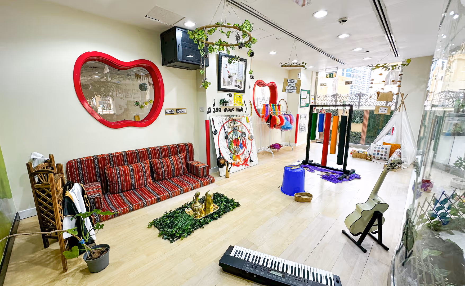 Bright indoor play area with music wall, colorful costumes, large xylophone tubes, a keyboard, and guitar on stands.