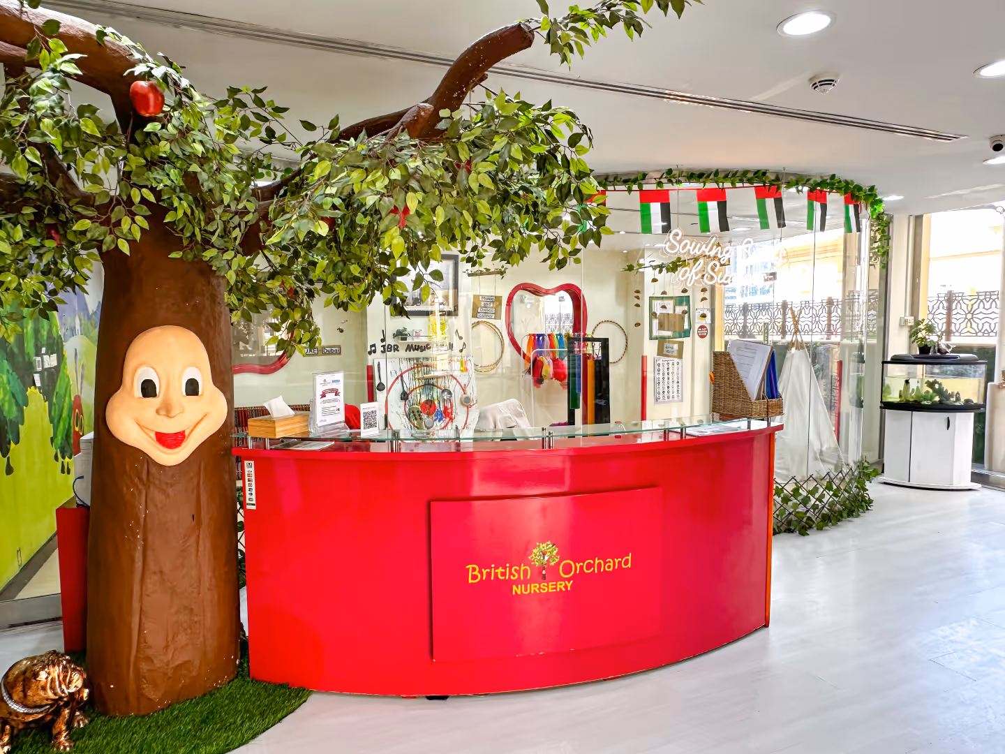 Reception area of British Orchard Nursery with a bright red desk, a tree structure with a smiling face, and UAE flags hanging above.