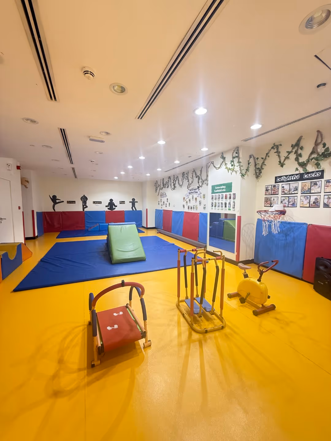 Indoor children's playroom with yellow floor, blue padded mats, small exercise equipment, a mini basketball hoop, and decorative greenery on walls.