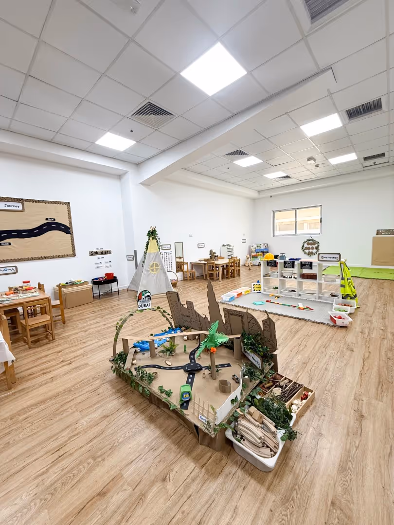 Bright classroom with wooden floor featuring a cardboard city model, small tables with chairs, a teepee, and labeled learning stations.