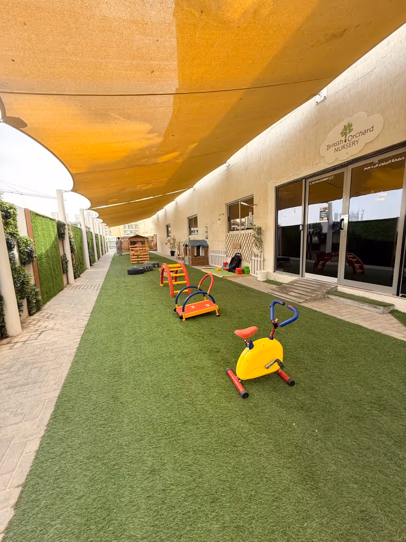 Outdoor play area at British Orchard Nursery with artificial grass, a yellow exercise bike, colorful play structures, and shaded canopy.