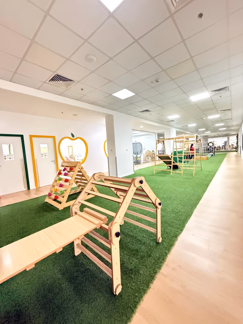 Indoor play area with green artificial grass floor and wooden climbing structures and ramps along a wide hallway.