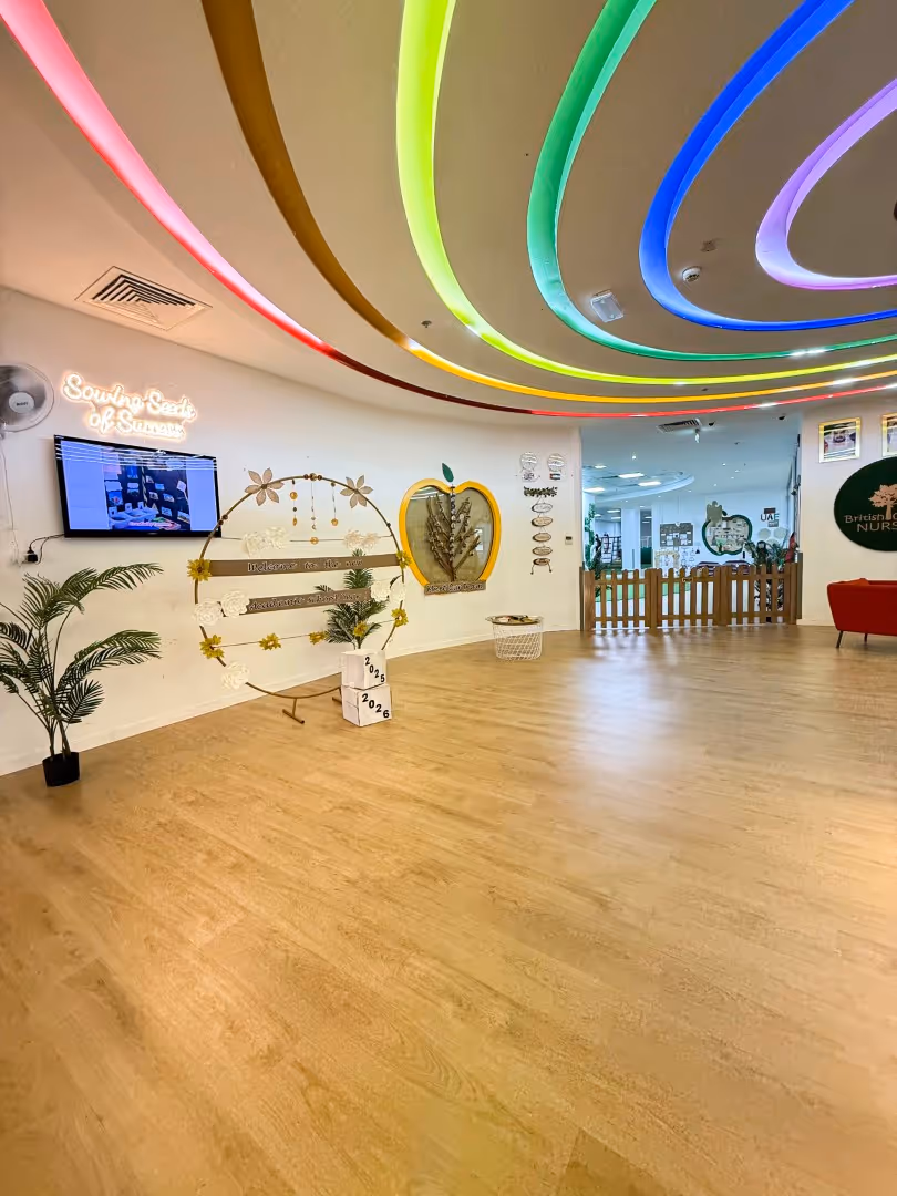 Bright room with wooden floor, colorful curved ceiling lights, a TV on the wall, a decorated circular display, and a plant in a pot.