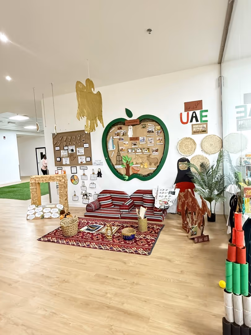 Classroom corner decorated with UAE cultural displays including a striped sofa, a camel cutout, traditional crafts, a green apple-shaped board, and UAE letters on the wall.