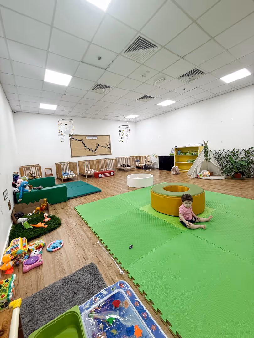 Spacious daycare room with a toddler sitting on a green play mat, surrounded by cribs, toys, and a small play tent.
