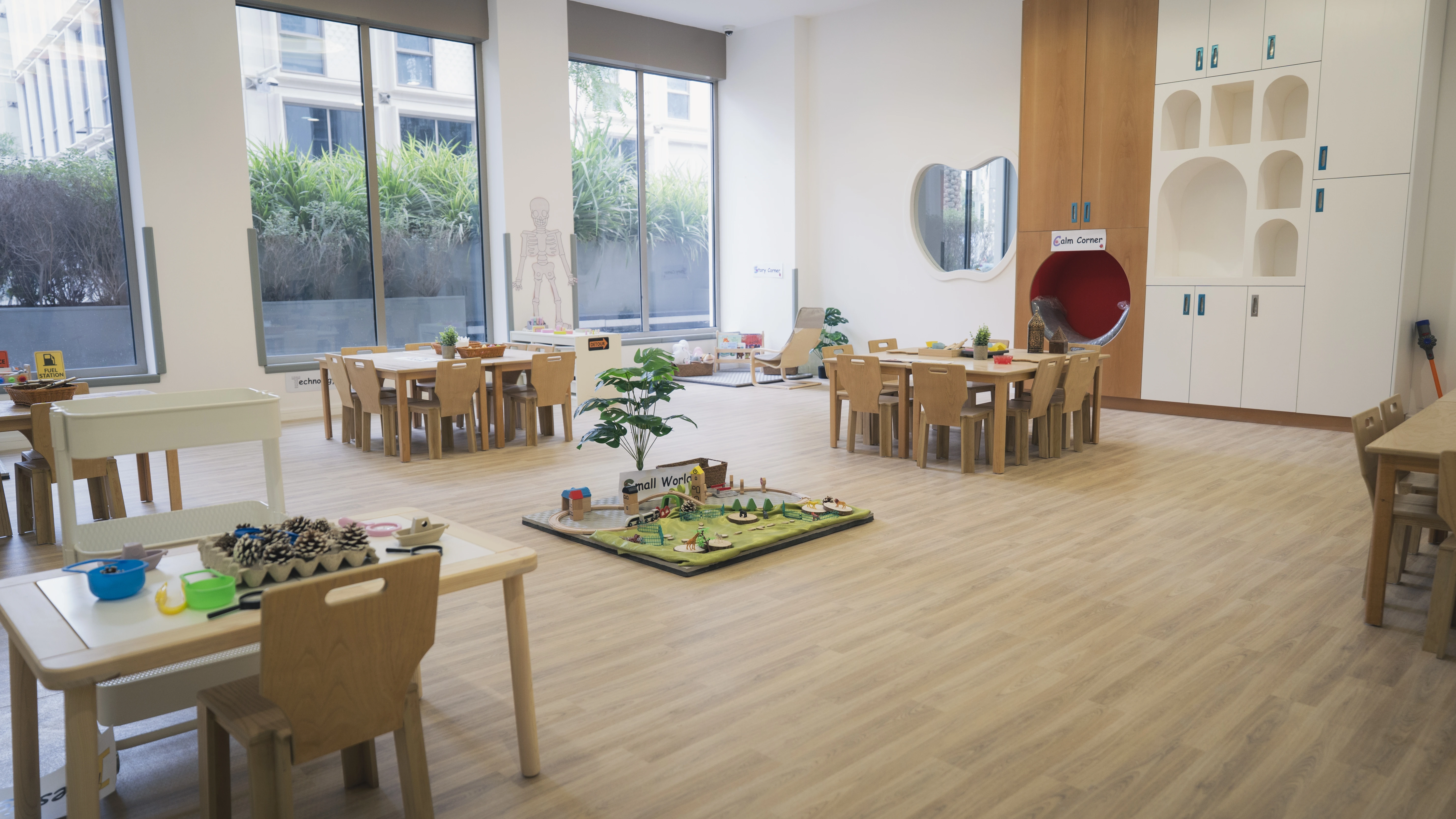 Bright, spacious preschool classroom with wooden tables and chairs, large windows, play area with toys, and a cozy Calm Corner with a round red seat.