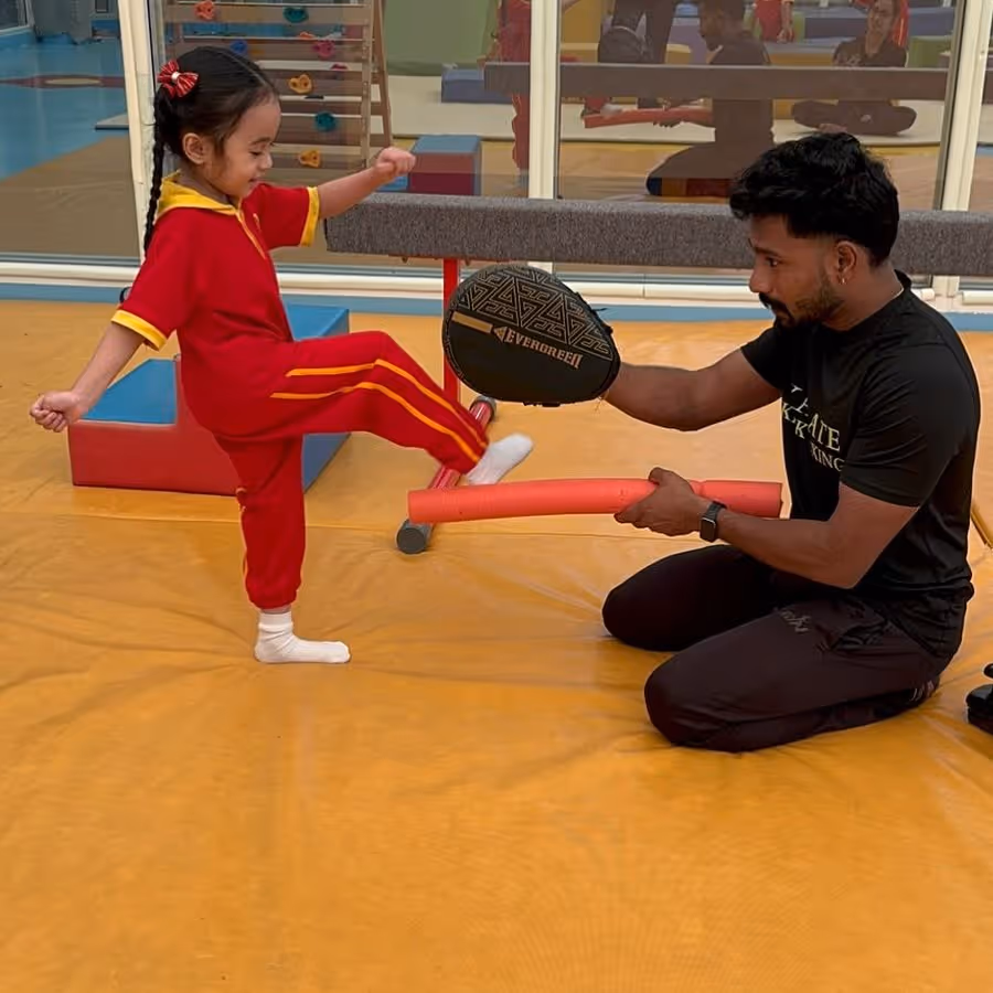 A young girl in red sportswear practices kicking with an instructor holding a pad and a foam stick in a gym.