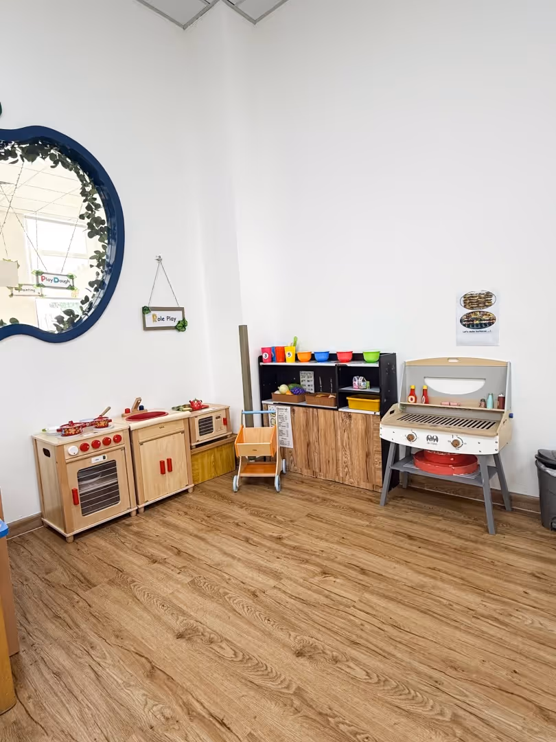 Children's play kitchen area with wooden toy stove, microwave, shelves with colorful bowls, toy grill, and a small wooden cart on a wood floor.