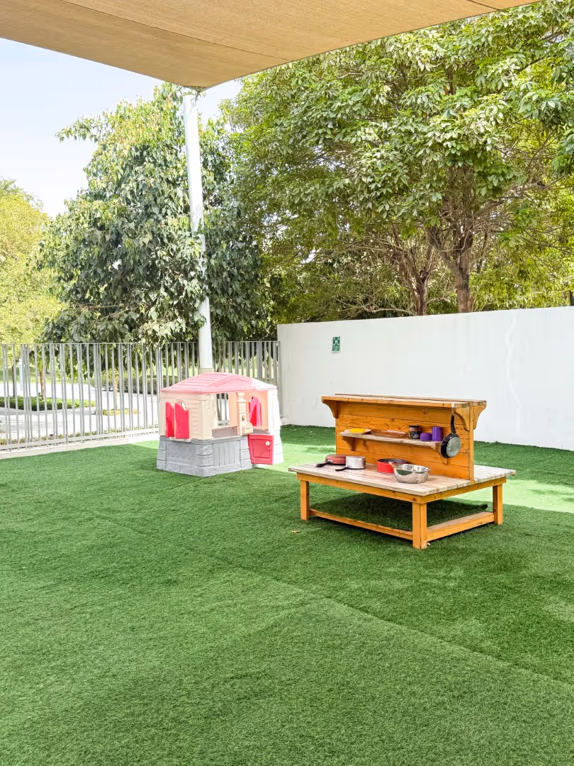 Outdoor play area with artificial grass, a small plastic playhouse, and a wooden play kitchen setup with toy utensils and pots.