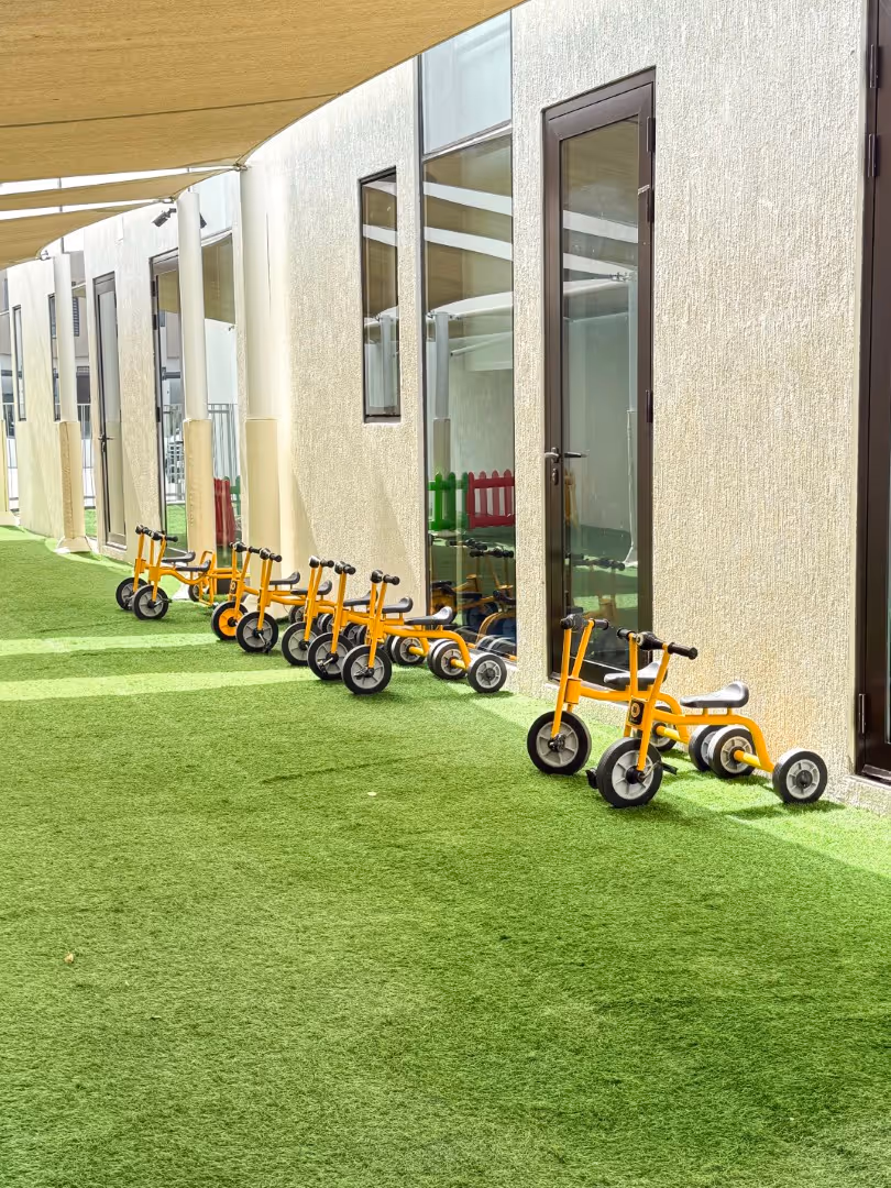 Row of small yellow tricycles lined up against a beige building wall on green artificial grass under a shade canopy.
