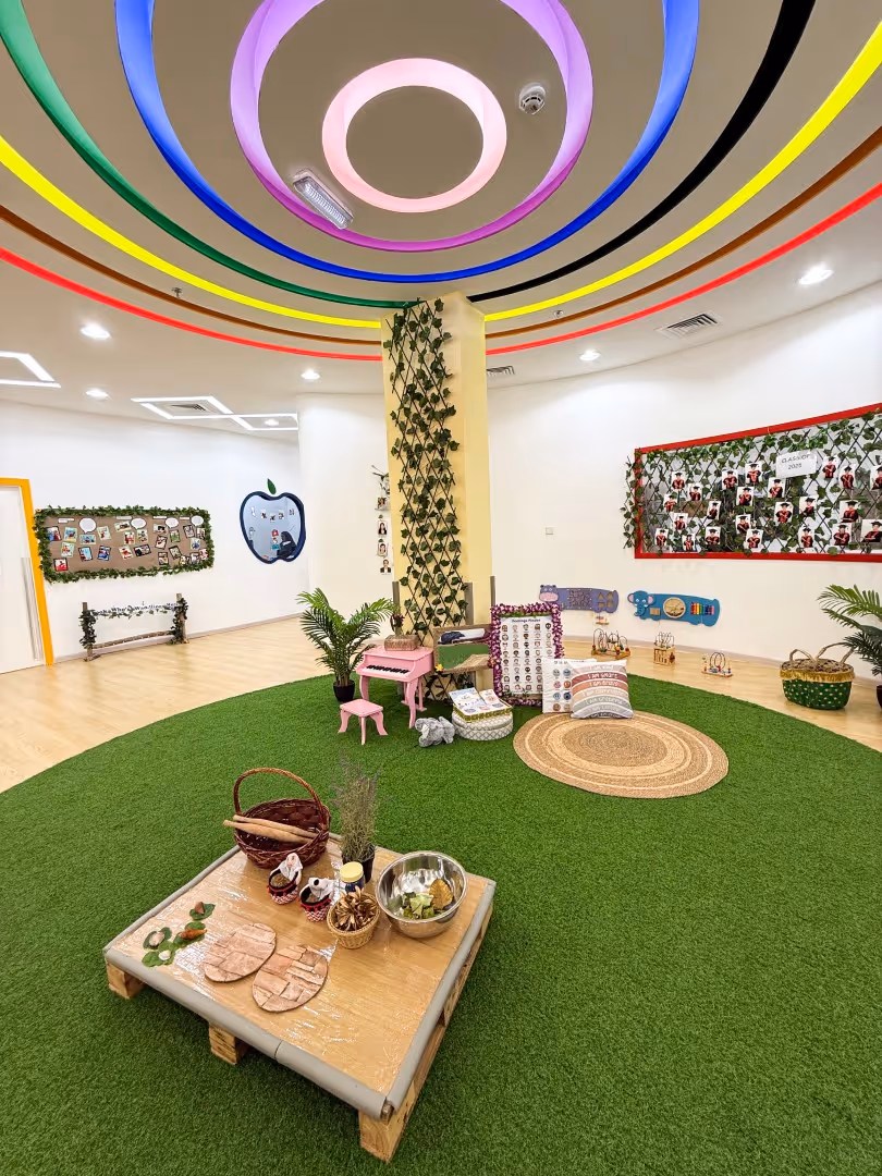 Bright indoor play area with artificial green grass, colorful circular ceiling lights, plants, toys, and educational wall displays.