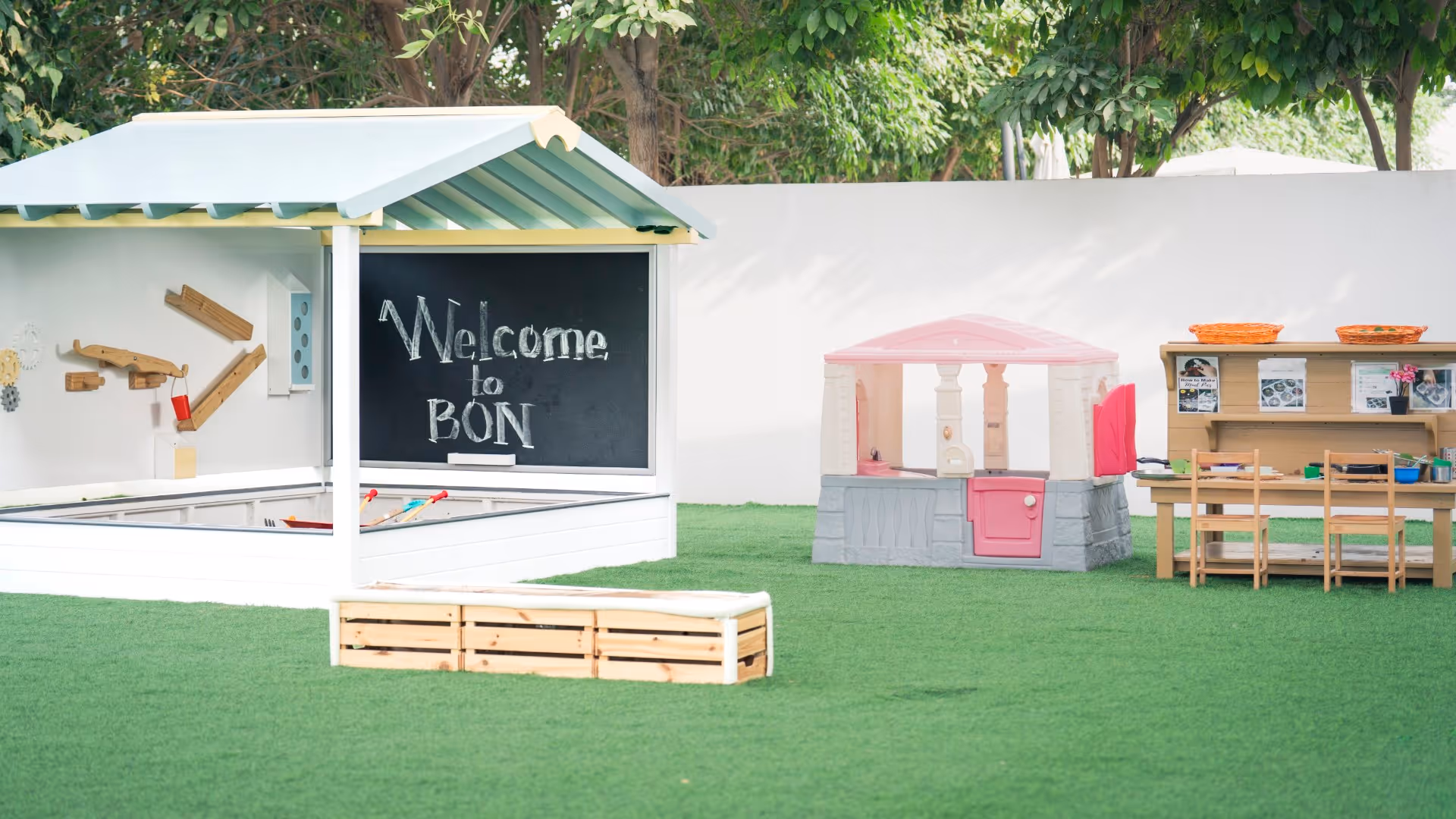 Outdoor play area with a small wooden structure, a chalkboard reading 'Welcome to BON', a pink and gray playhouse, and a wooden table with chairs on green artificial grass.
