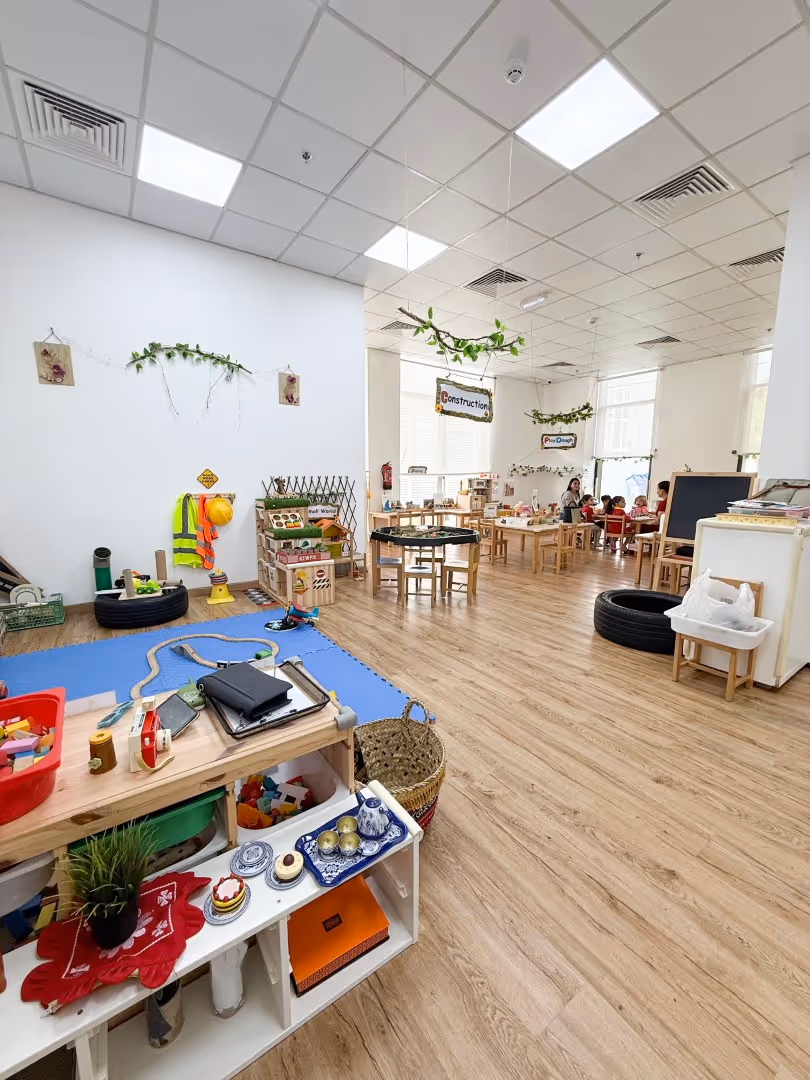 Bright and spacious classroom with play areas, small tables, chairs, toys, and children engaged in activities under signs labeled Construction and Play Dough.