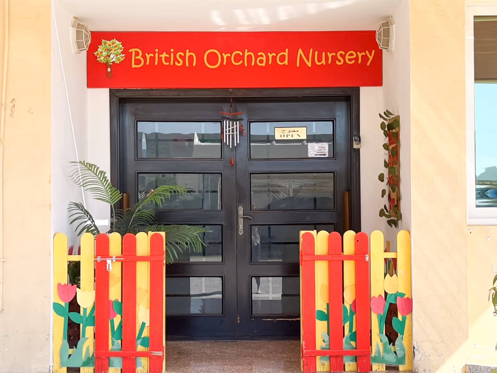 Entrance to British Orchard Nursery with a black door, a red sign above, and colorful red and yellow picket fences decorated with tulip designs.