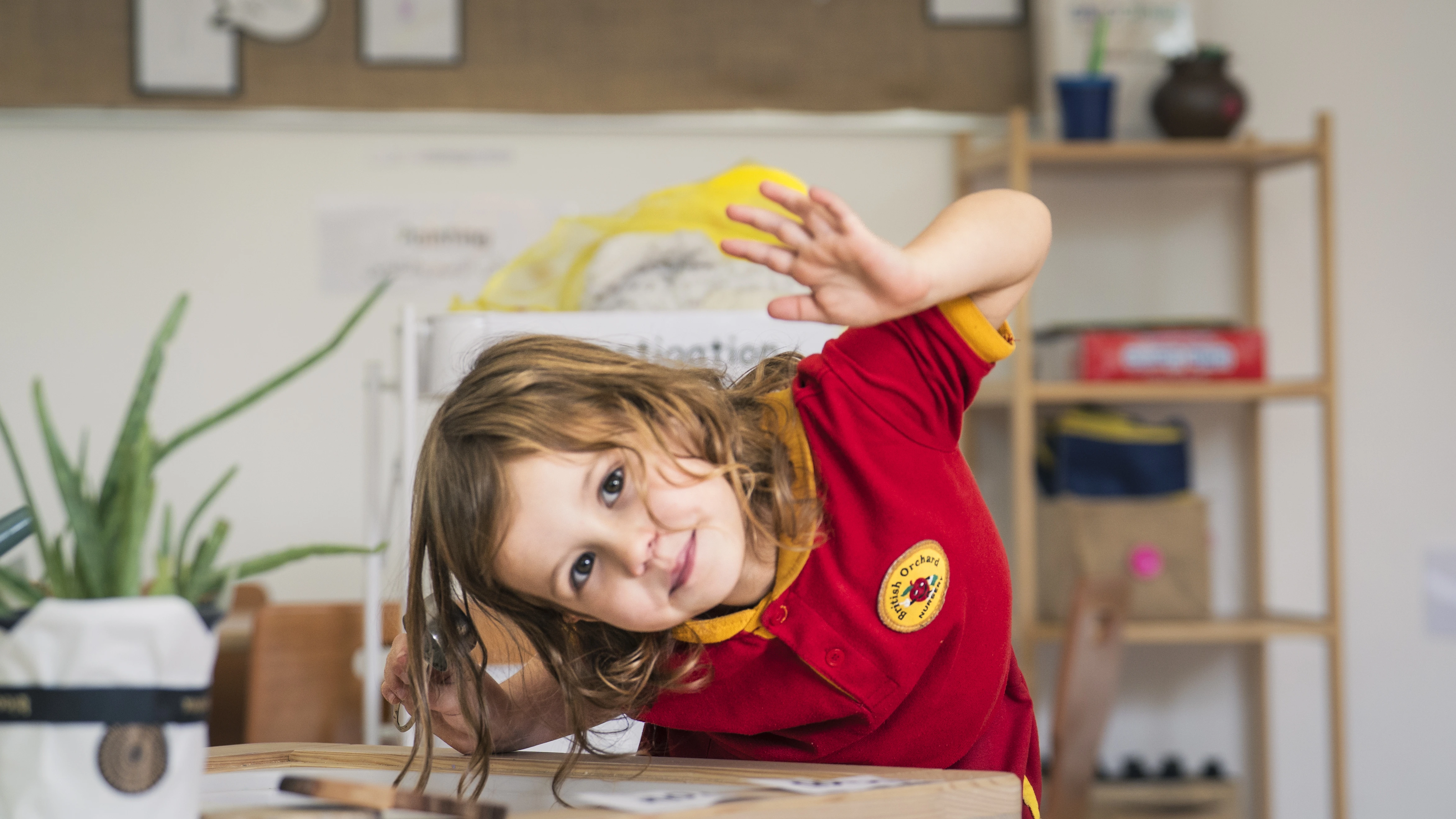 Young child in a red school uniform leaning sideways while playing at a wooden table in a classroom.
