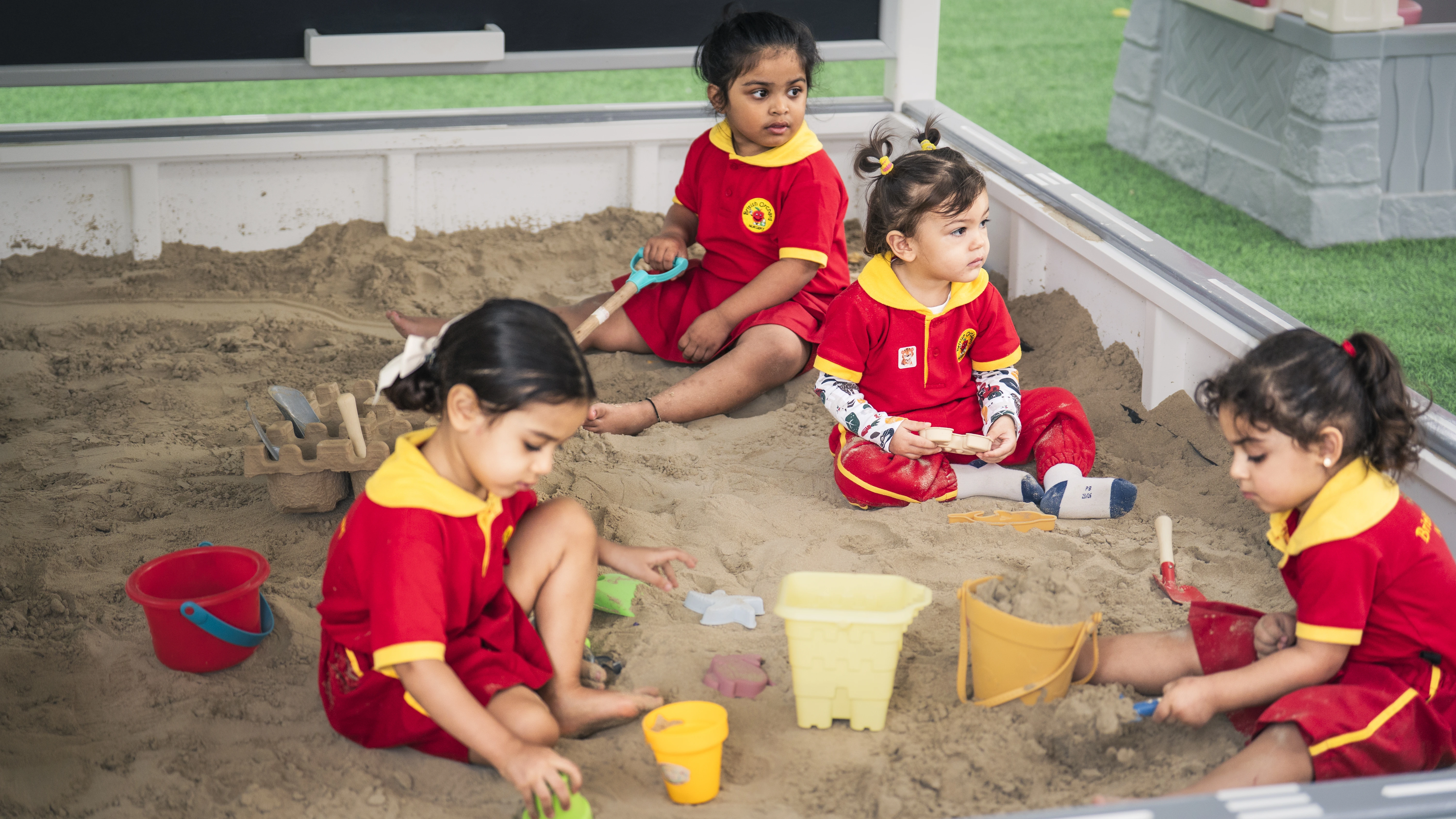 Four young children in red and yellow uniforms playing with sand and toys in a sandbox.