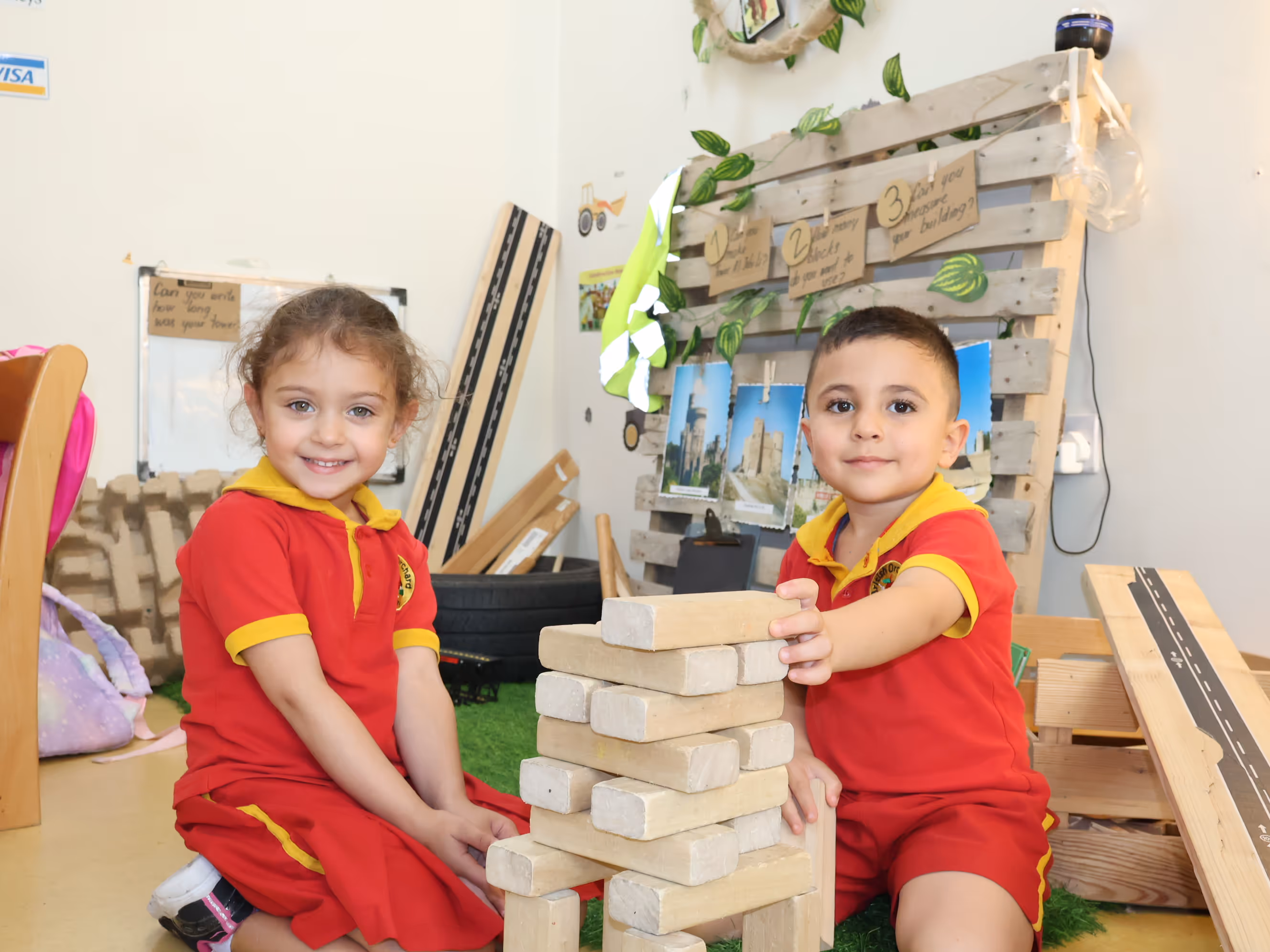Two young children in red and yellow uniforms smiling while playing with wooden block tower indoors.