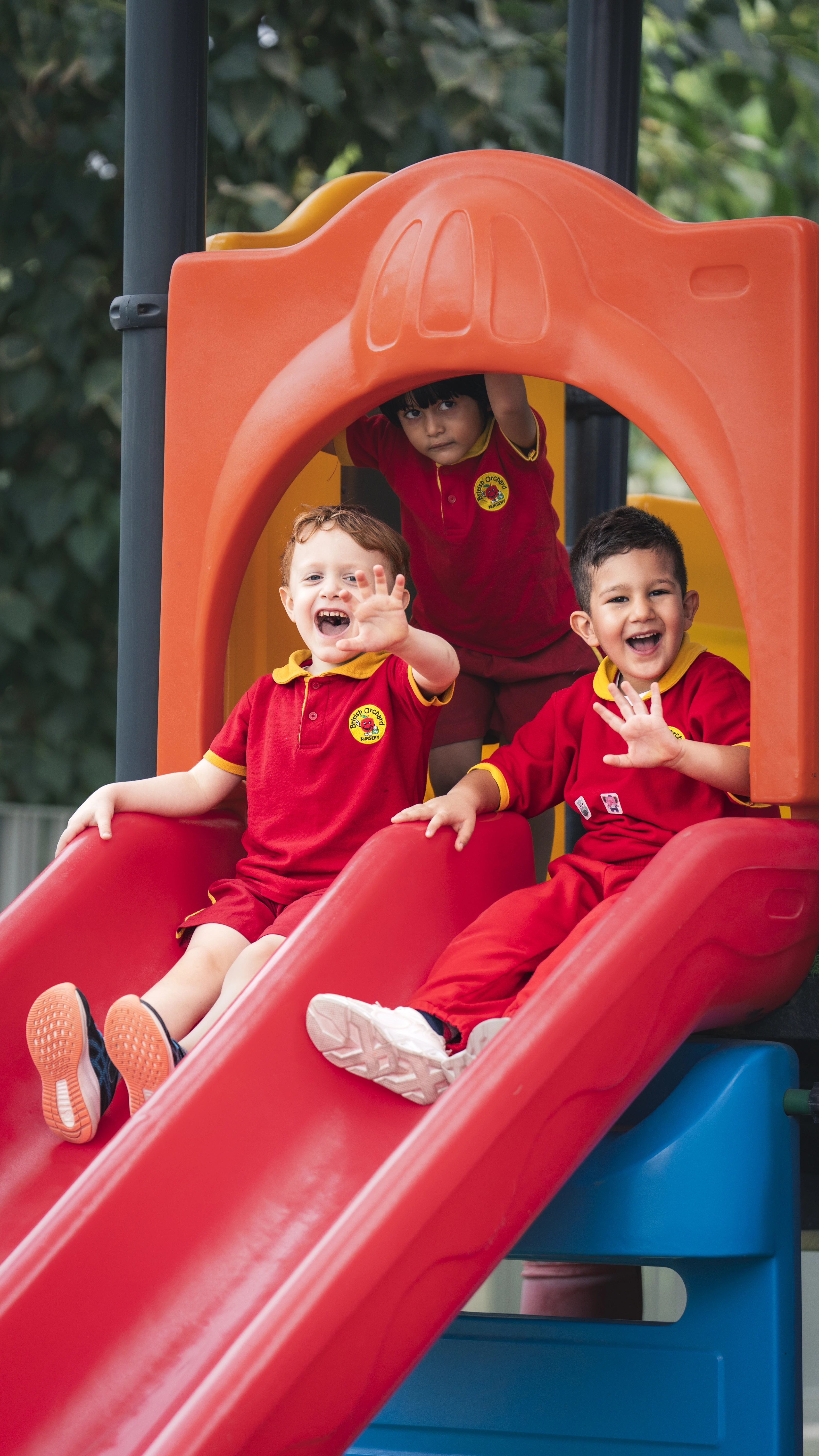 Three children in red nursery uniforms playing on a red double slide, two sliding down and one standing behind them.