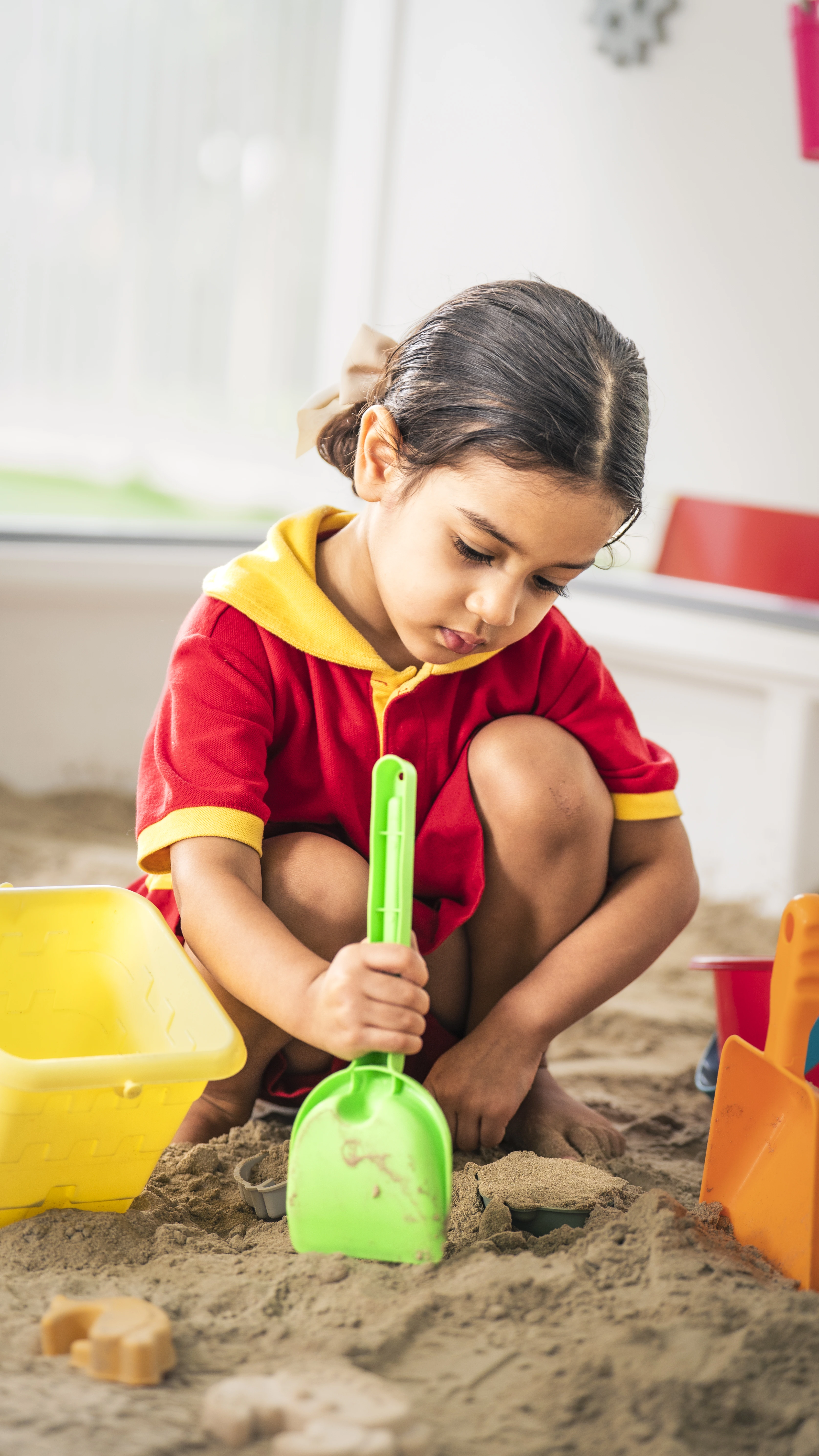 Child in a red and yellow outfit playing with a green shovel in indoor sandbox.