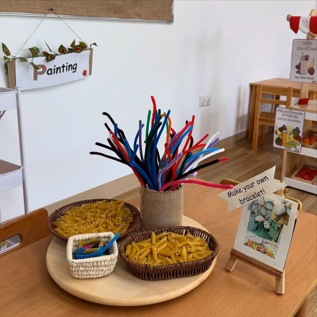 Table with baskets of uncooked pasta and colorful pipe cleaners for making bracelets, next to a sign that says 'Make your own bracelet!'