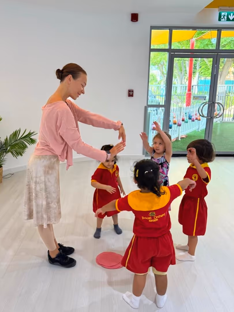Teacher leading a group of young children in an indoor activity, raising their arms, with three kids in red and yellow nursery uniforms.