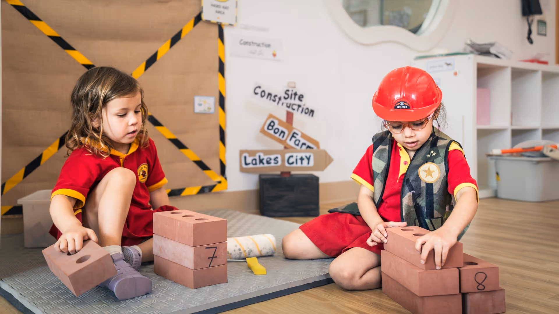 Two children playing with toy bricks, one wearing a red hard hat, in a classroom with a construction-themed setup.
