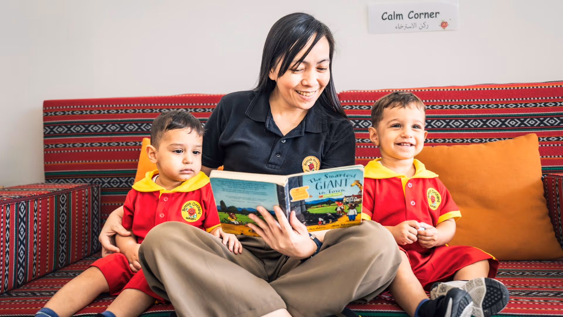 Woman reading a book to two young boys sitting on a patterned red couch in a room labeled Calm Corner.